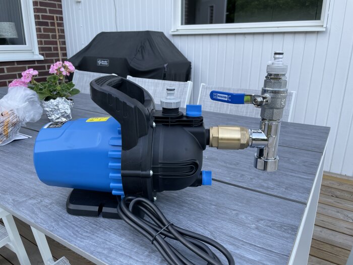 Blå vattenpump på träbord, grill överdrag, blomkruka, folieinslagna föremål i bakgrunden.