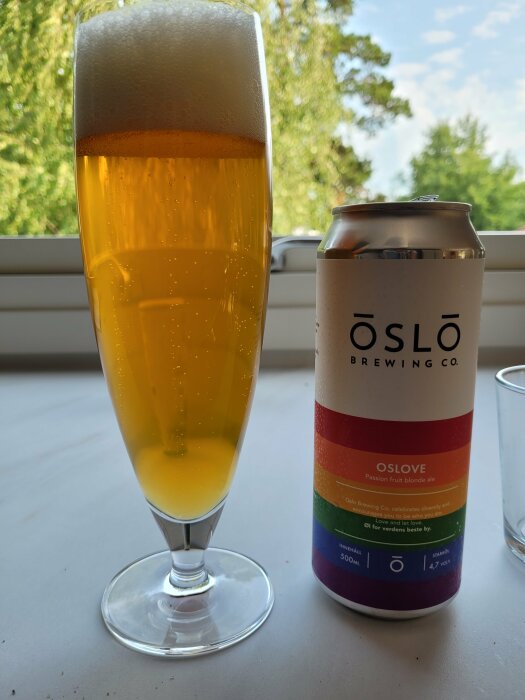 Ett glas öl med skum vid ett fönster och en ölburk från Oslo Brewing Co.