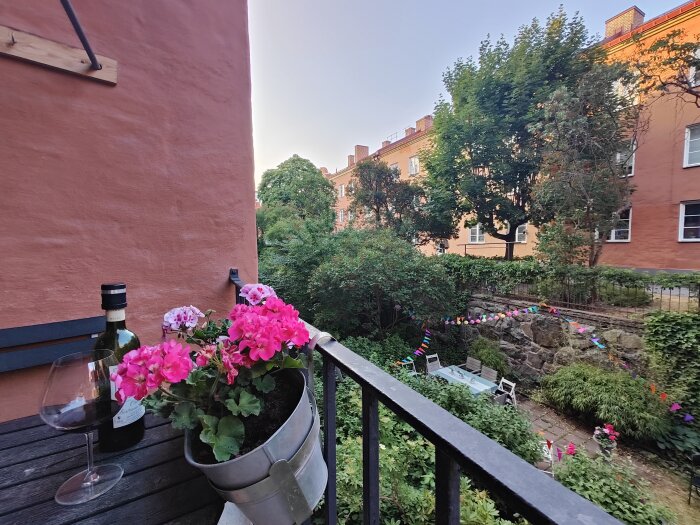 Balkongvy med rosa blommor, vinflaska, glas, innergård och bostadsbyggnader under kvällsljus.