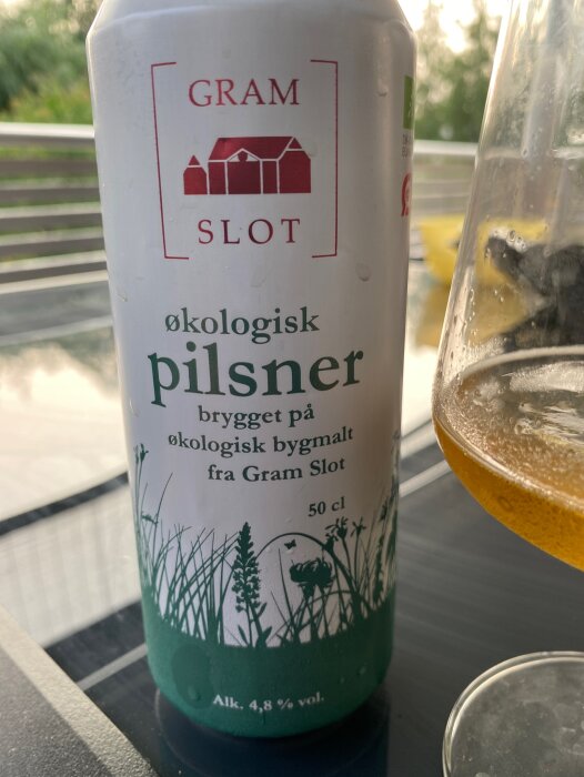 En ölburk av märket Gram Slot, ekologisk pilsner, på ett bord nära glas.