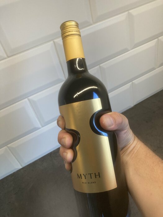 En hand håller en flaska rödvin med etiketten "MYTH Red Blend" framför en vit vägg.