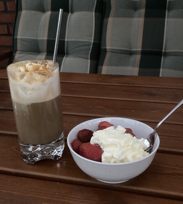 Kall kaffe med grädde och metall sugrör. Jordgubbar och grädde i skål. Träbord. Rutig bakgrund.