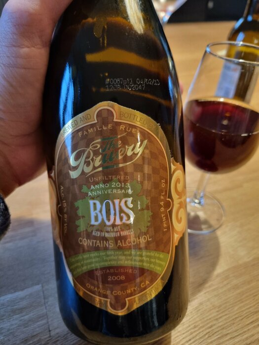 Ölflaska hålls, etikett visar "The Bruery Bois", årgång 2013, bourbonfatlagrad ale, bakgrund med vinglas.