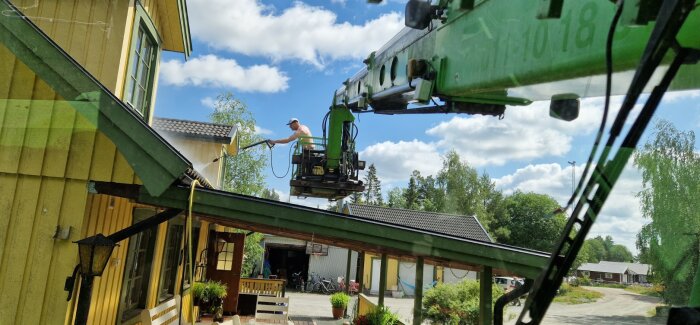 En man använder en höglyftande kran för att tvätta ett gult trähus utomhus på en solig dag.