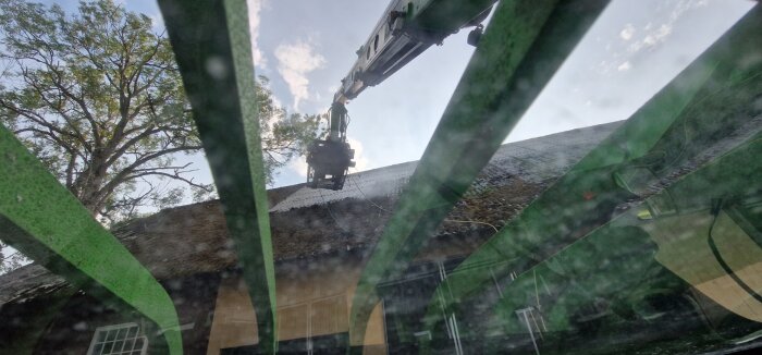 Genom smutsigt fönster ses bomlift vid gammalt hus med stråtak, träd i bakgrunden, himmel, reflektioner.