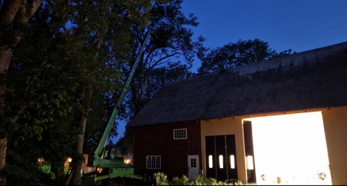 Kväll, röd ladugård, öppen dörr med starkt ljus, lyftkran, träd, delvis molnig himmel.