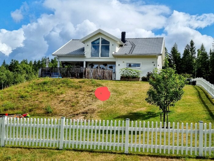 Vit villa med veranda på kulle, gräs, träd, vit staket, molnig himmel.