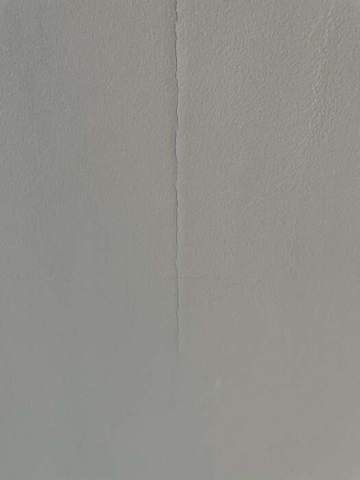 Enkel vit vägg med en vertikal spricka, ojämn yta, skuggor, ingen kontext.