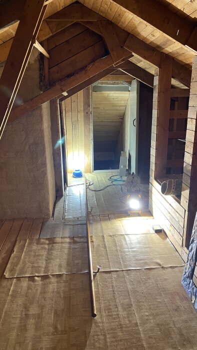 Renoveringsarbete i vindsvåning med belysning, isolering, träkonstruktion och kablar. Oinredd, byggarbetsplats-stämning, fokus på struktur och ljus.