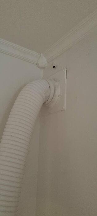 Flexibel vit ventilationsrör ledas genom en vägguttag i ett hörn av ett rum.