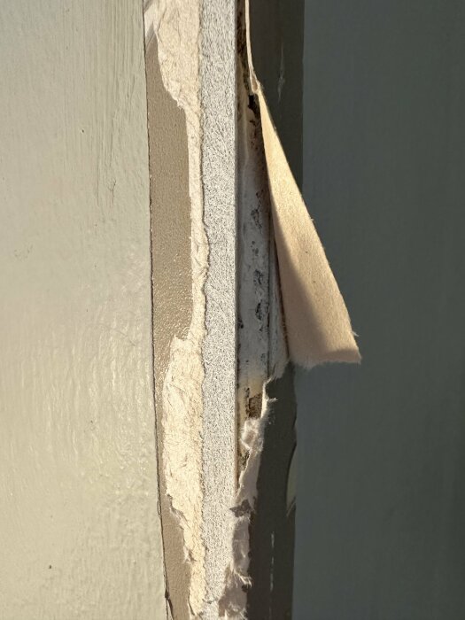 Skadad vägg med flagande målning och tapet som avslöjar flera lager i solljus.