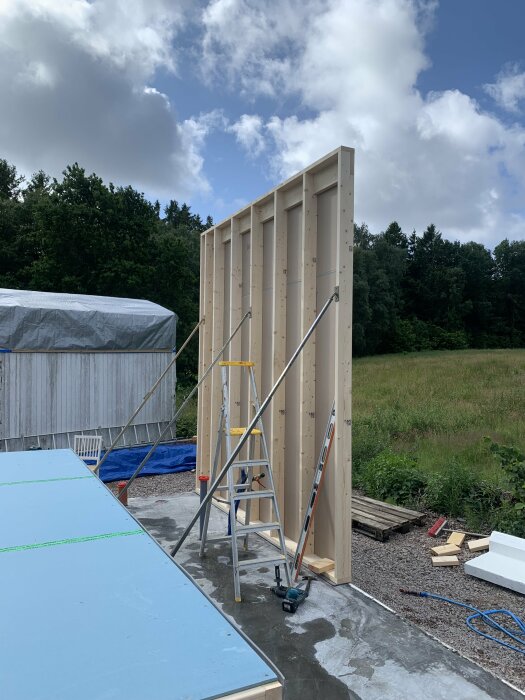 Byggplats med stående träväggkonstruktion, stegar, verktyg och material på grusunderlag under molnig himmel.