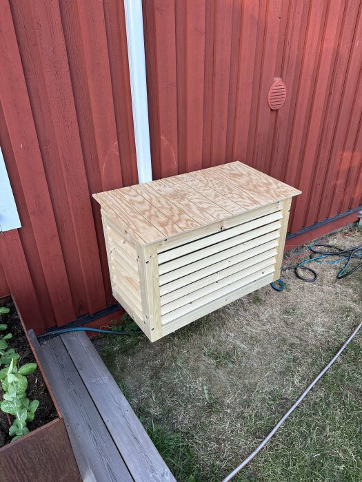 Träkompostbehållare i trädgård mot röd stuga, gräsmatta, ventilationsgaller på vägg.