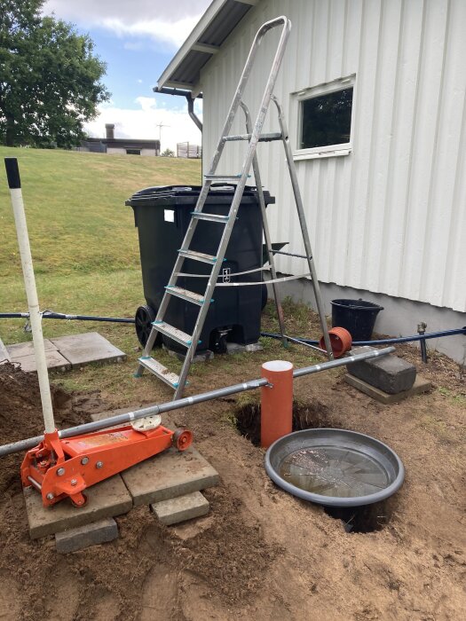 Grävd grop, stege, hydraulisk domkraft, rör, sopkärl, gräsmatta och husvägg. Arbetsplats eller reparation utomhus.