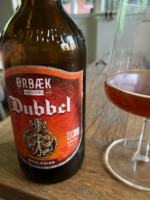 Ölflaska med etikett "Ørbæk Dubbel", närbild, delvis fullt glas, ekologisk öl, inomhus, träyta, skandinavisk design.