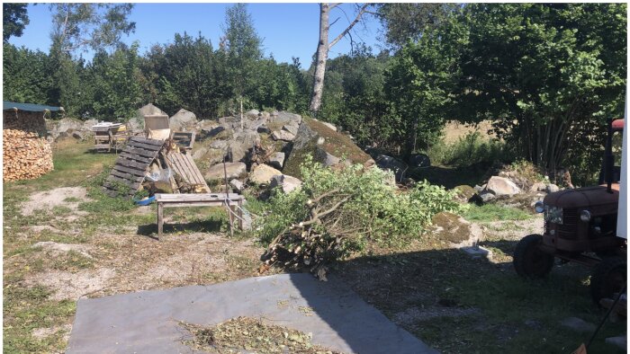 Lantgårdsmiljö med vedhög, stenar, traktor, nedfallna trädgrenar och landbruksutrustning på en solig dag.