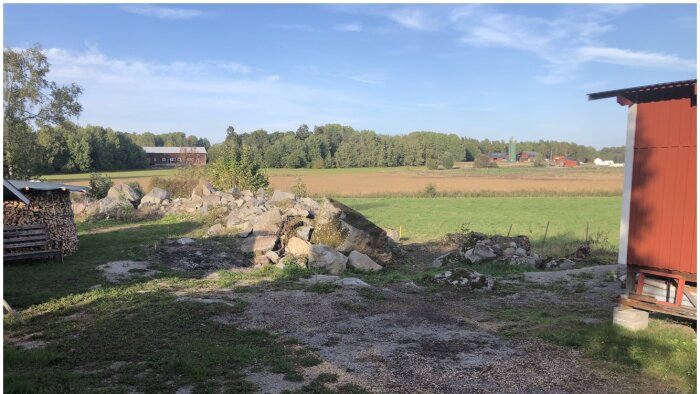 Lantlig vy med vedhög, stenröse, gårdar, åkrar och klar himmel.