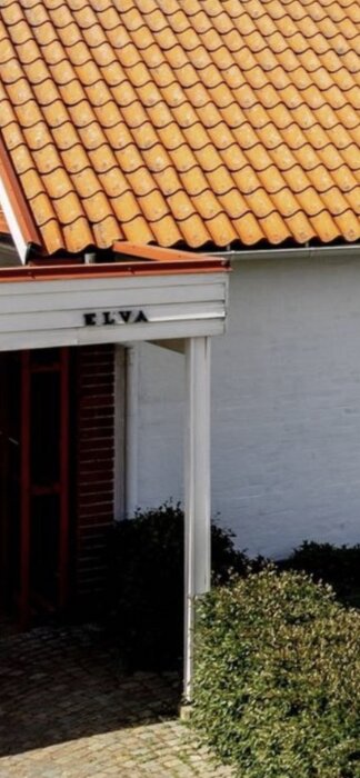 Ett vitt hus med tegeltak, träfönsterluckor, skylt som säger "ELVA", buskar och en stenlagd gång.