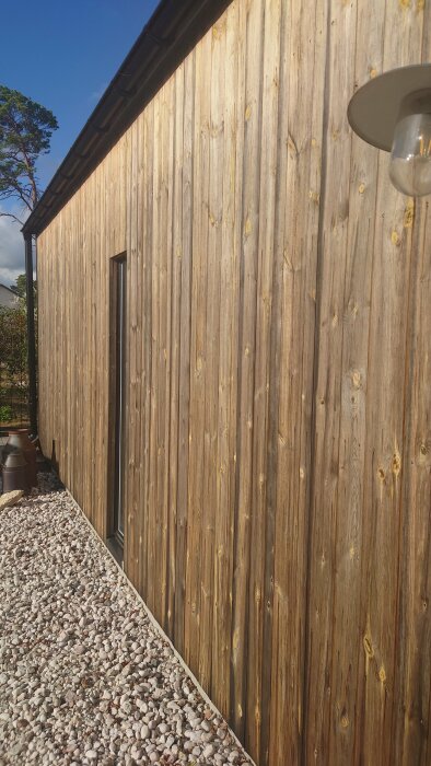 Träfasad på hus, fönster, utomhuslampa, grusväg, grönska, blå himmel, dagsljus, arkitektur, modern, minimalistisk design.