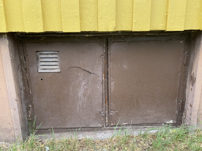 Brun ventilation, metallucka, gul vägg, slitet, gräs, ingen synlig handtag, utomhus, krypgrundsentre.