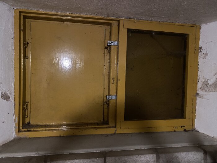 Slitet gult väggskåp med öppen dörr, på en rå betonghylla, gammalt och nött, i dunkel miljö.