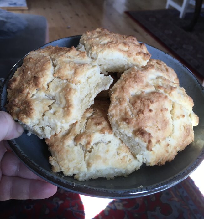En person håller en tallrik med nybakade, fluffiga scones. Hemtrevlig interiör i bakgrunden.