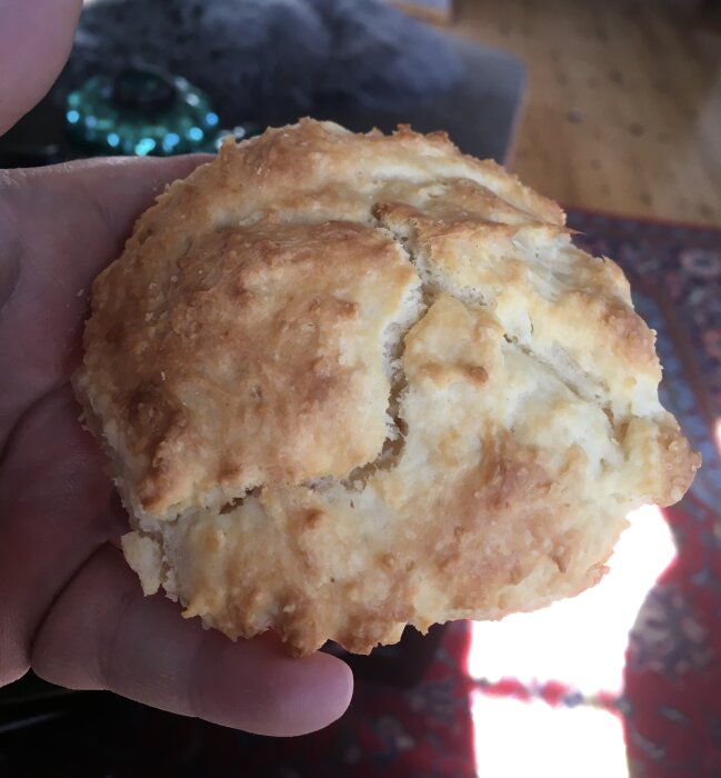 Någons hand håller en nybakad, gyllenbrun scones. Bakgrunden är suddig med orientalisk matta.