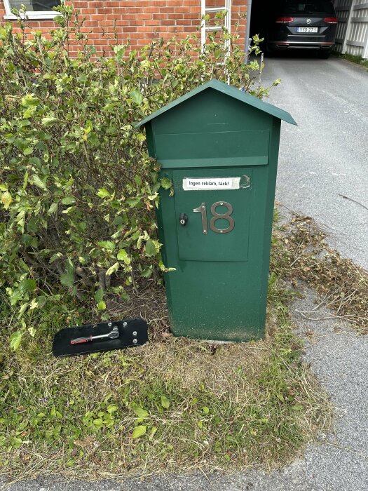 Grön brevlåda med nummer 18 och skylt "Ingen reklam, tack!", vid trottoar, nära buskage och bakom bil.