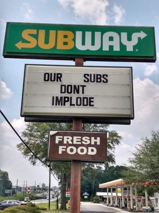 Skylt: "SUBWAY" logotyp. Nedan, budskap: "OUR SUBS DONT IMPLODE". "FRESH FOOD" nedanför. Dagtid, molnig himmel, bakgrund med mack.