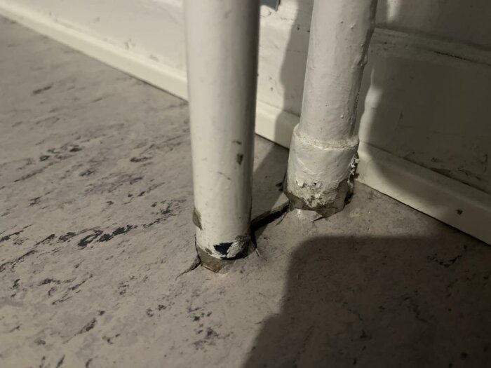 Två vita rör går ned i sprucken grå betonggolv, nära vit list, slitage och skador synliga.
