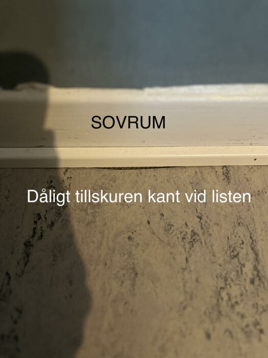 Vit dörrkarm med texten "SOVRUM", skugga, fokus på dåligt avskuren golvlist.
