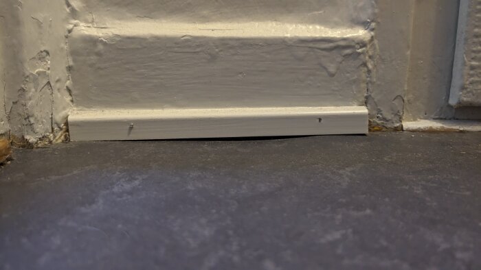 Golv möter vit dörrfoder vid texturerad vägg; slitet, ojämnt, visar ålderstecken och behov av underhåll.