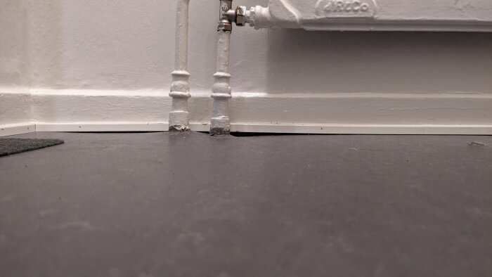 Vitmålade rör och en radiatorventil mot en vägg över ett grått golv.