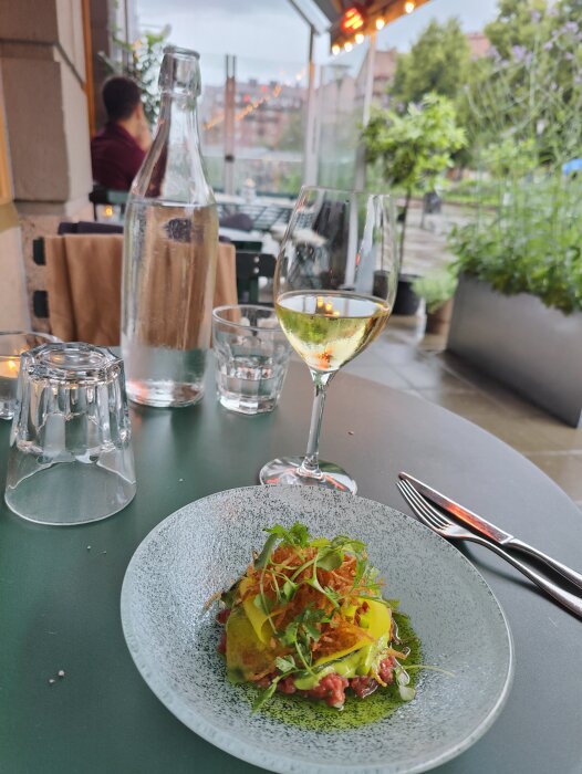 Uteservering med maträtt, vitt vin och vattenflaska. Grönskande utsikt och suddig person i bakgrunden.