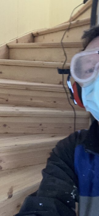 Person med andningsskydd och skyddsglasögon framför trätrappa. Spegling, otydlig, suddig. Arbetsklädsel synlig.