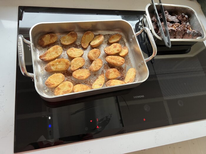 Stekta halverade potatisar och brownies i metallformar på en induktionshäll. Köksscenario, matlagning, bakning, rostade potatisar.