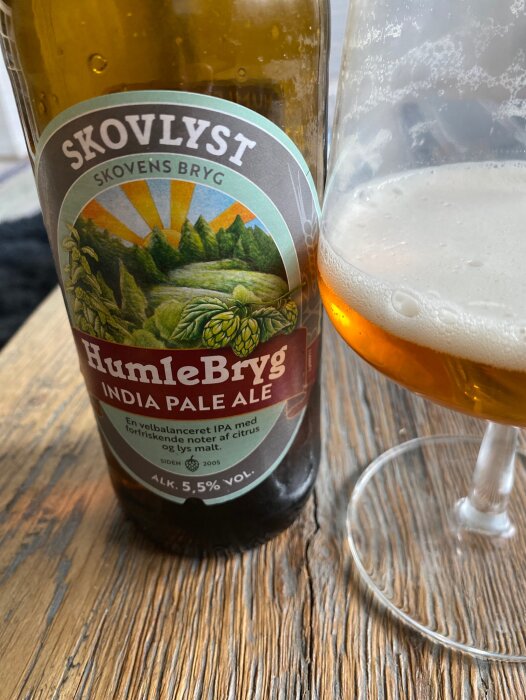 Ölflaska märkt "Skovlyst" och glas med öl. Etikett med skog och humle. India Pale Ale. Träbord.