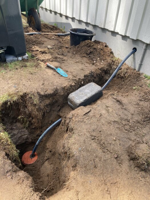 Grävd grop i jord nära byggnad, spadar och skottkärra synliga. Arbete eller reparation kan pågå.