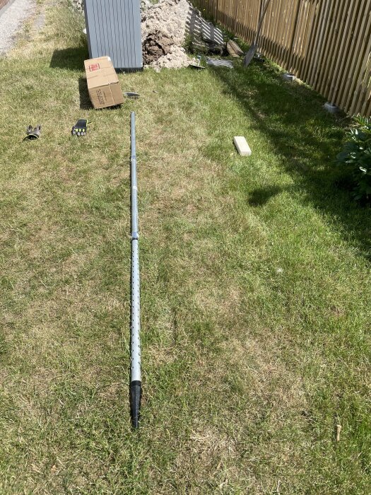 Lång metallstång ligger på gräs. Kasserad förpackning, trädgårdshandskar, träbit, hög med jord bakom staket.