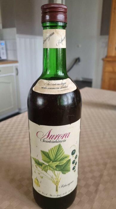 Flaska Aurora Svartvinbärssaft, grön etikett, försluten, står på bord, diffus bakgrund, köksmiljö.