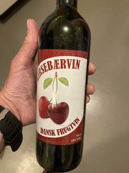 Hand håller en flaska dansk fruktvin med körsbärsetikett och texten "Kirsebærvine". Armbandsur syns.