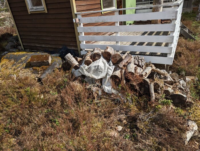 Hög med ved och presenning bredvid trähus och vit trästaket. Naturlig, solig dag.