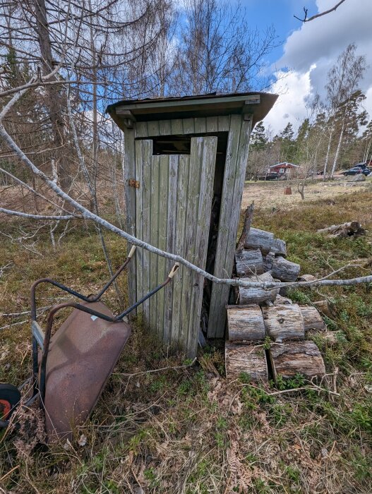 Förfallen trähus, utedass, rostig skottkärra, uppstaplade stockar, natur, delvis molnig himmel, lantlig miljö.