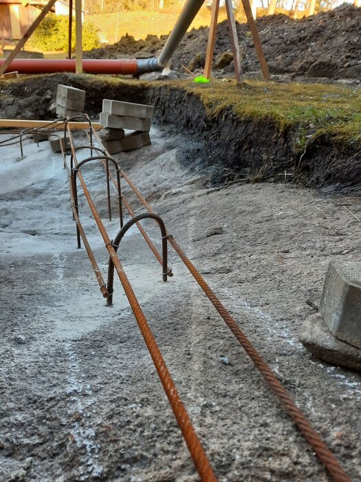 Byggarbetsplats med armeringsjärn, betongplintar och rörsystem på en grusig mark.