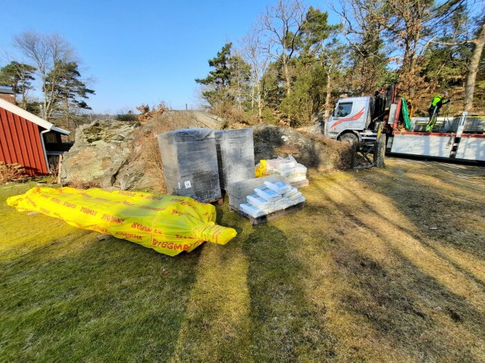 Leverans av byggmaterial, säckar och stenull, med lastbil och kran, på en trädgård nära skog.