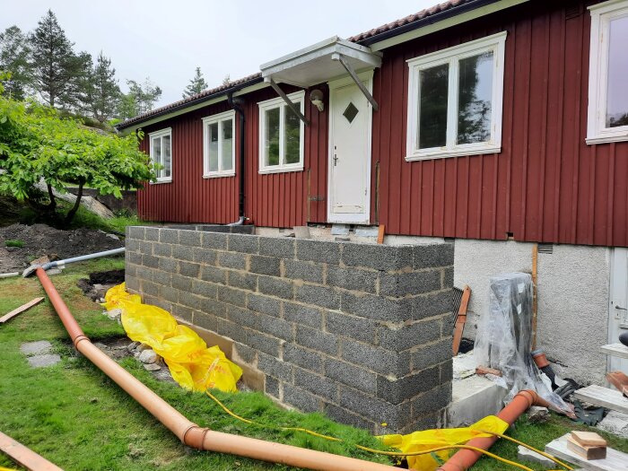 Röd trähusfasad, grå grundmur under konstruktion, byggmaterial och rör utspridda, gråmulen dag.