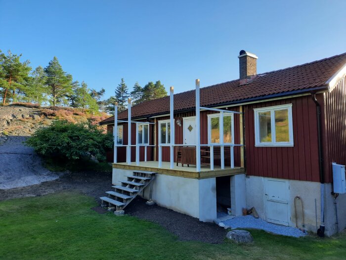 Rött hus med vita knutar, veranda, grönt gräs, träd och klippor under blå himmel.