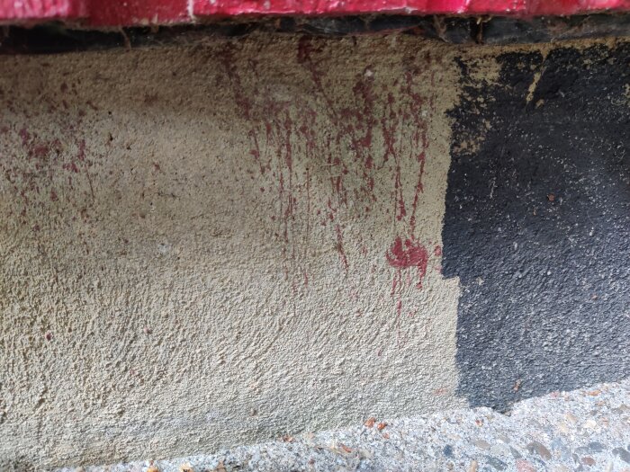 Röd färg droppar på betongvägg, under röd kant, bredvid svart målning. Slitage och smuts synligt.