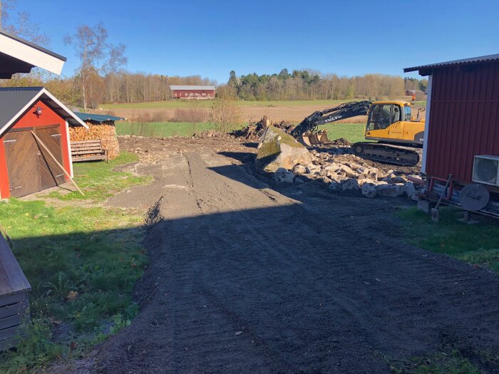 Grusväg, grävmaskin, nedrivna byggnadsrester, ladugård i bakgrunden, soligt vädret, utomhusscen, ingen person synlig.