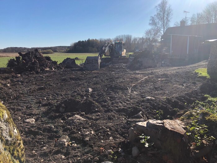 Grävmaskin på markarbetsplats, trädrot, jordhögar, klart väder, delvis synligt hus.